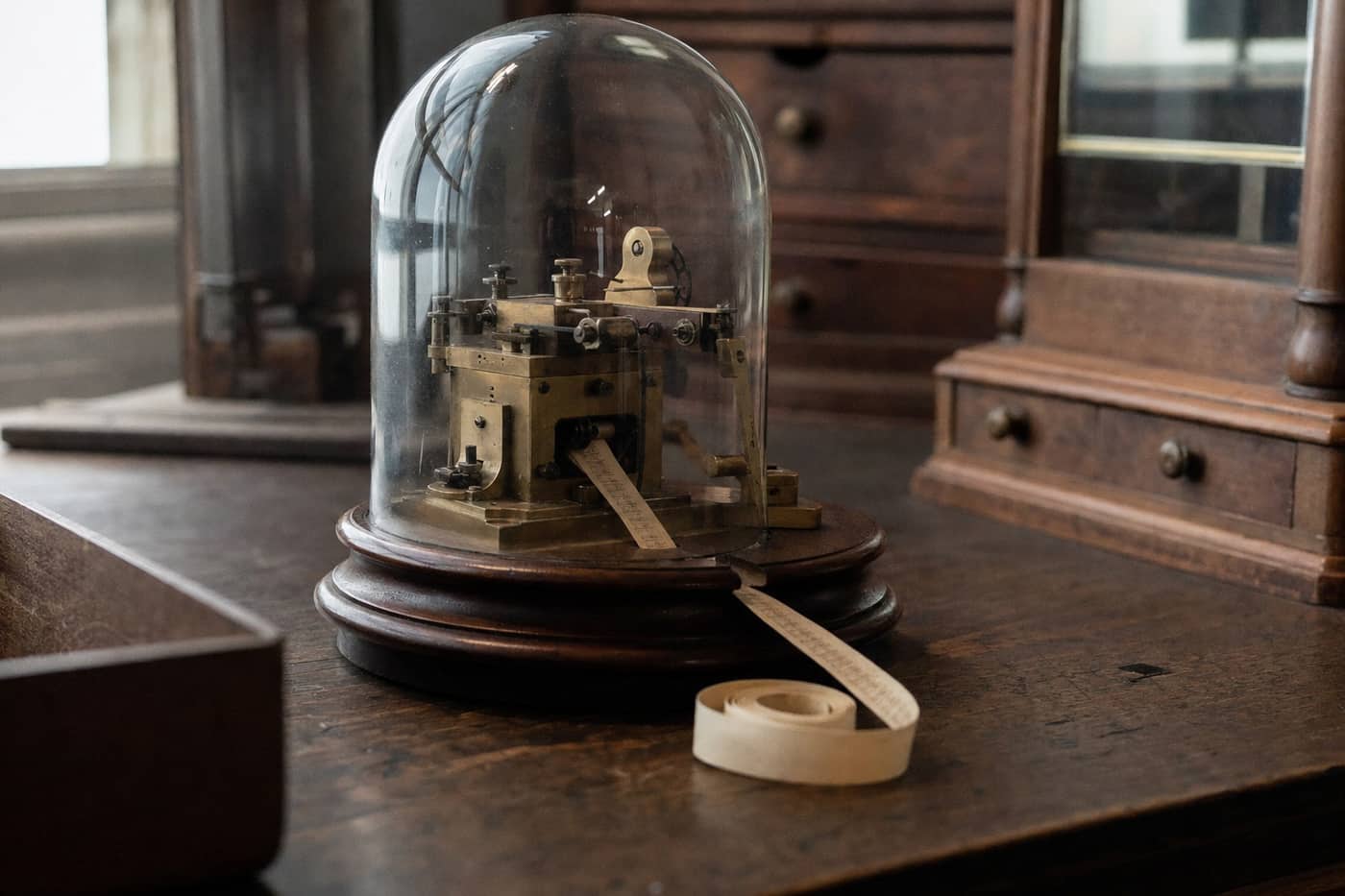A nineteenth-century stock ticker under a glass dome, its paper tape coiling on the desk.